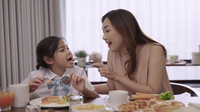 Happy Asian family mother and child daughter in a school uniform sitting smiling and eating breakfast for health together before school on the dining table during morning in the kitchen room at home. 