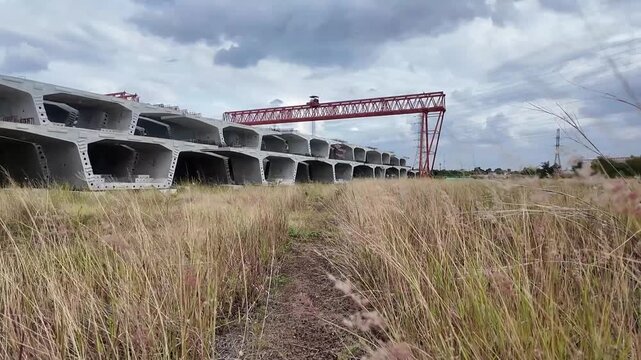Massive precast concrete bridge segments are neatly stacked in a vast, overgrown grassy field under a cloudy sky.