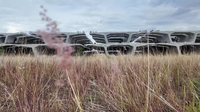Massive precast concrete bridge segments are neatly stacked in a vast, overgrown grassy field under a cloudy sky.