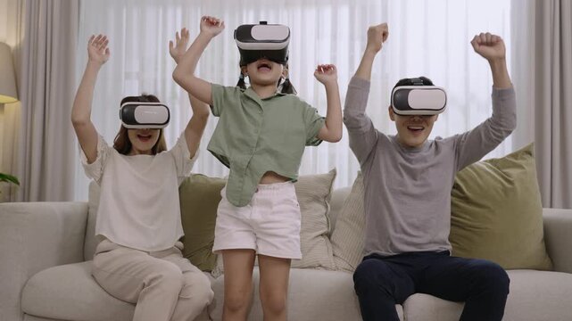 A moment of happy Asian family father, mother, and daughter wearing VR 3d or virtual reality glasses, playing video game together on the sofa in the living room at home. Relaxing innovation concept. 