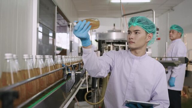 Asian quality control inspector in a uniform food and drink factory holding a tablet inspecting fruit juice bottles while moving on conveyor belt in beverage production line at manufacturing industry.