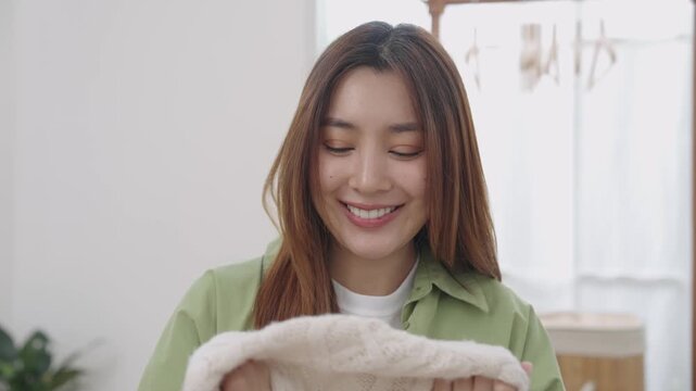 Happy, serene Asian woman holding and smelling freshly washed cream knit sweater with eyes closed, smile in bright cozy laundry room at home