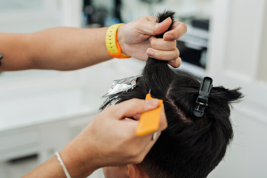 Hair stylist sectioning hair and placing foils for highlights during professional hair highlighting treatment for an Southeast Asian man in his early 20s