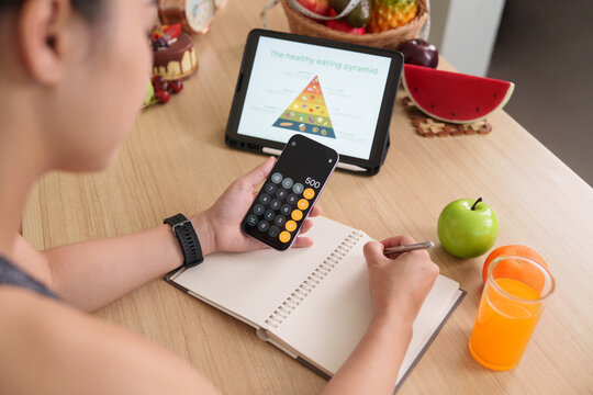 Woman writing in notebook while calculating calories with phone for healthy eating pyramid meal plan