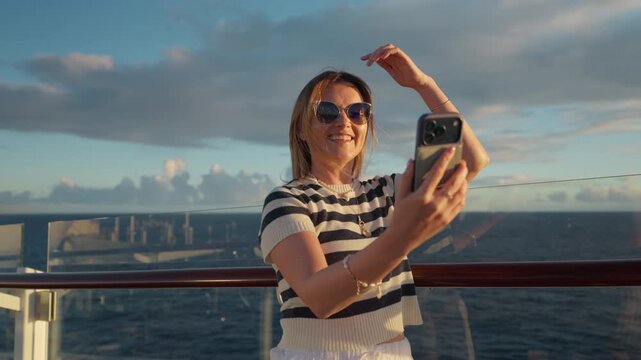 Smiling woman wearing striped nautical top and sunglasses video calls on smartphone while standing at cruise ship railing during golden hour with dramatic clouds and calm ocean waters in background.
