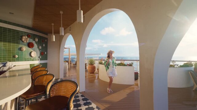 Stylish woman in straw hat walks through elegant cruise liner outdoor bar lounge in Caribbean Sea. White arches and Mediterranean decor frame ocean views as golden sunlight illuminates relaxing deck.