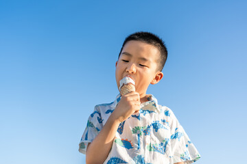 A young asian boy enjoys eating ice cream outdoors against a clear bright blue sky. © tai