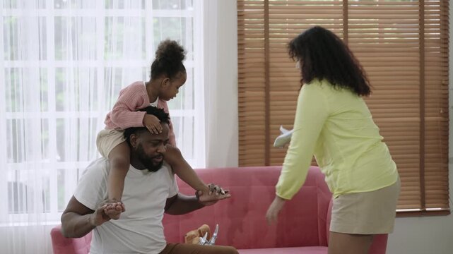 Happy African family. A cute daughter rides on her father's shoulders, holding and playing with a plane model with her mother, having fun and cheerful together on the sofa in the living room at home. 