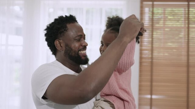 Happy African American father standing smiling and holding his cute little daughter while dancing together with love and bonding, having fun and cheerful in living room at home. Father's Day concept. 
