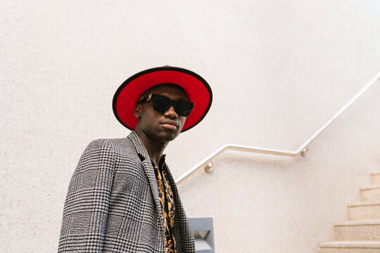 Fashionable man wearing hat and sunglasses standing in front of wall