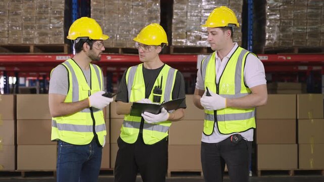 Teamwork of three warehouse workers in uniform wearing yellow safety helmets, standing and working talking and discussing planning in large logistics distribution factory. Inventory stock concept. 