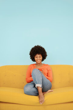 Happy woman using mobile phone sitting on yellow sofa against blue background
