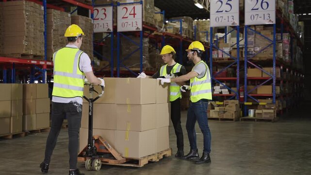 Warehouse workers in yellow safety helmets, standing and working, talking and discussing planning and moving box cargo by hand pallet truck in logistics distribution factory. Inventory stock concept. 
