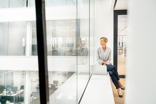 Blond businesswoman with laptop, sitting on windowsill in office building