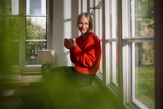 Smiling mature woman sits by the window holding a mug at home