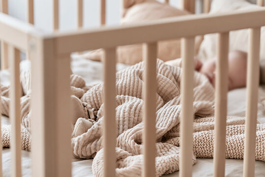 Softly blurred view of a Scandinavian nursery crib with a sleeping infant's legs covered in a textured blanket, warm morning light illuminating the scene