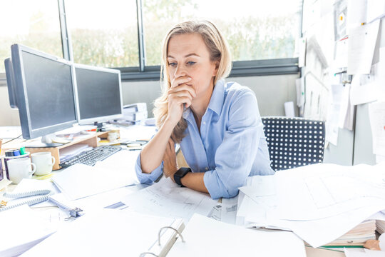 Pensive woman sitting at desk in office surrounded by paperwork