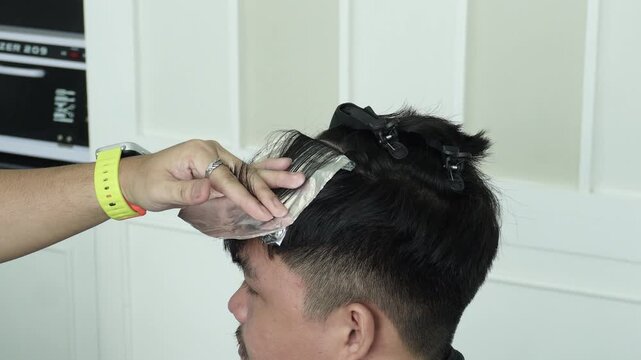 Hair stylist applying lightener to hair on foil during professional foil highlights treatment for Southeast Asian man in his early 20s