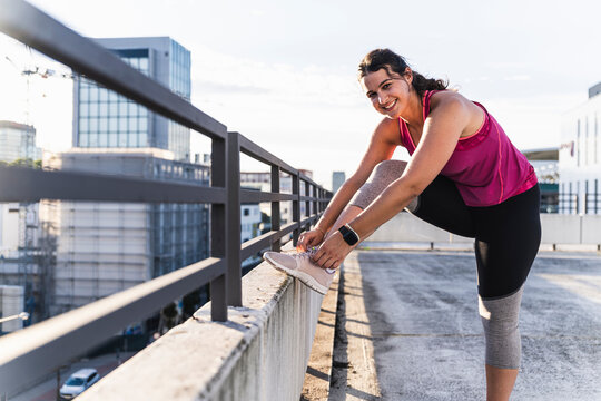 Smiling young woman tying shoelace on railing against sky