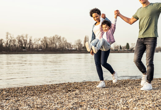 Cheerful boy swinging with parents support over pebble by lake