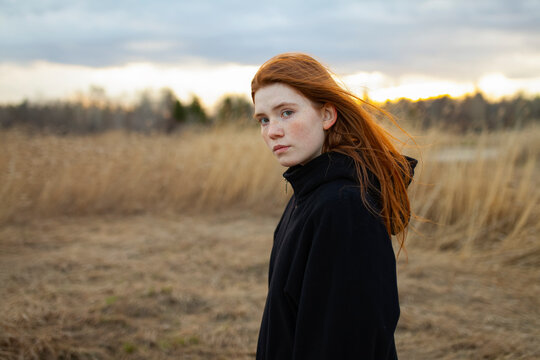 Portrait of teenage girl with red hair in reed field wearing black hoodie at sunset