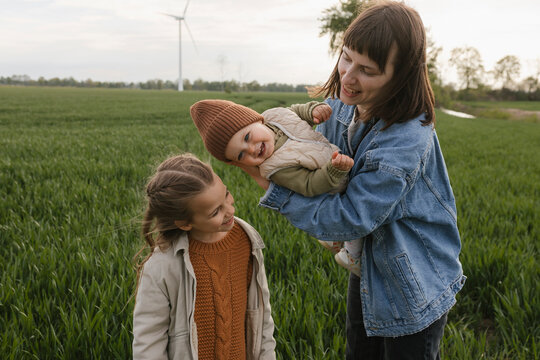 Mother holds baby beside daughter in green field with wind turbine