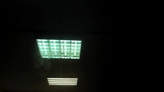 Ceiling light fixture emitting smoke in a darkened room. Fluorescent lamps flickering and releasing vapor in an office interior. Safety hazard and fire concept.