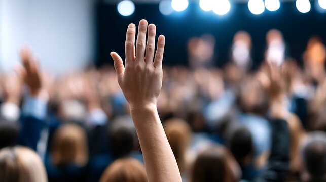 Person raising a hand in a blurred audience at a public event, engaging in active participation and interaction during a seminar or presentation