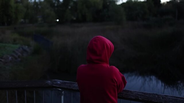 A woman in a red hoodie stands contemplatively on a wooden bridge near water at dusk, eyes closed in prayer. Soft natural evening light, outdoor park setting, reflective mood captured.