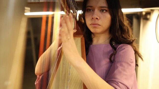 Close-up shot of a woman in a mauve dress expertly playing a harp, with emphasis on delicate finger movements, gleaming strings, and warm, soft-focused indoor studio lighting.