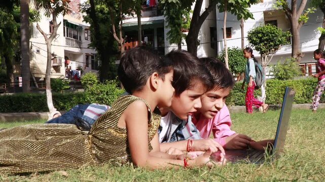 Elementary age cute loving siblings of Indian ethnicity lying down on grass in the park during springtime and using laptop together.