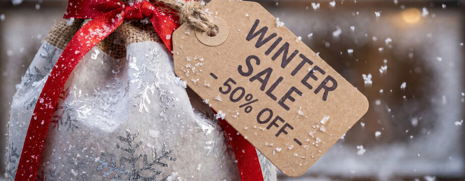 Burlap gift bag with winter sale tag and red ribbon. Christmas discount promotion with snow falling. Festive retail shopping concept during seasonal holiday clearance event.