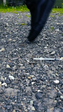 Close up view of a person wearing black shoes dropping a used hypodermic needle on the gravel ground and walking away, representing addiction, abuse, and the opioid crisis epidemic