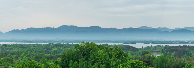 Obraz premium A wide landscape showing misty distant mountains, a calm lake, and dense green forest under an overcast sky.
