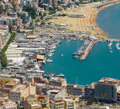 Aerial view of a sunny coastal town featuring a bustling marina filled with boats, turquoise sea waters, a sandy beach, and charming seaside buildings. It's Anzio port, in province of Rome, Italy.