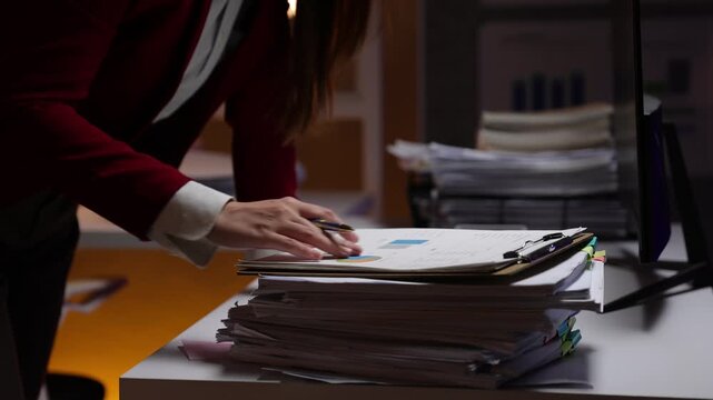 Businesswoman working late at night in a dark office, reviewing reports with charts and graphs on a large stack of documents.