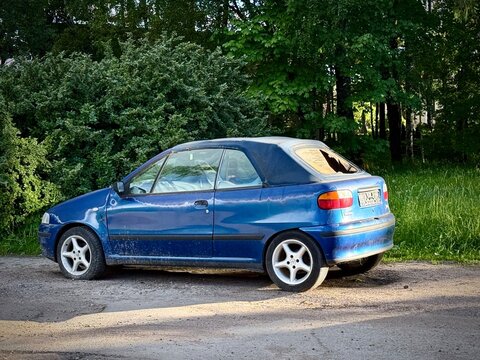 A first-generation Fiat Punto Cabrio (Type 176). The problem of abandoned cars in the city. A blue first-generation Fiat Punto Cabrio (Type 176) with a broken rear window is parked on a dirt road. 