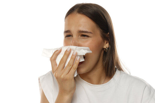 Portrait of a young woman sneezing into a tissue with closed eyes, on white background.