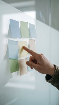 Hand selecting pastel sticky note on glass planning wall during brainstorm session