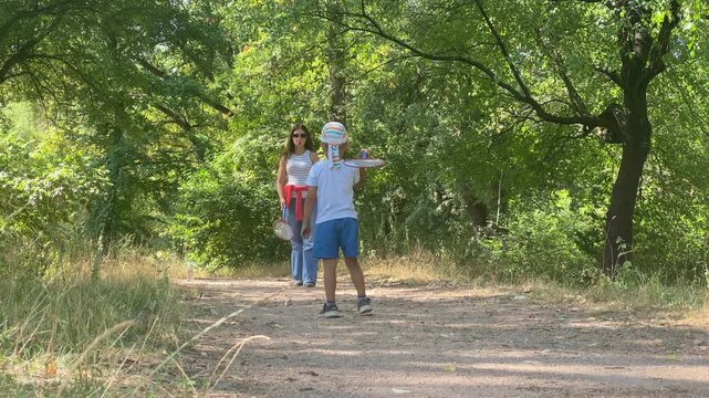 A mother and her little boy play badminton together. They hold rackets along a sunny path in a green forest. Their game creates a joyful and peaceful feeling.