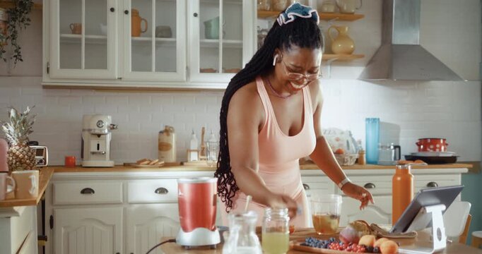 Smiling Black woman in sportswear playfully tosses fruit into a blender while making a healthy smoothie in her kitchen before a workout.