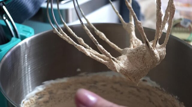 Whisk attachment lifted above stainless steel mixing bowl showing thick beige batter clinging to wires and forming peaks, batter strands stretching between wires and bowl rim indicating recent
