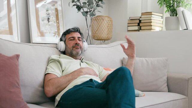 Video of mature man listening music with headphones while relaxing on couch at home