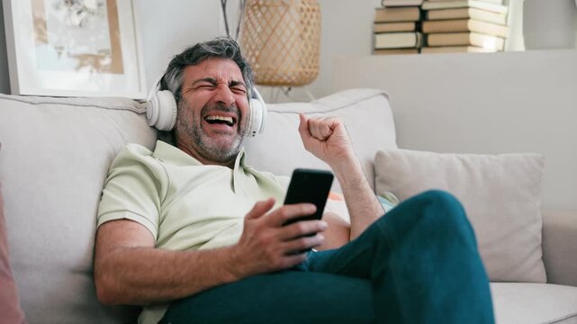 Video of mature man using smartphone while listening music with headphones on couch at home