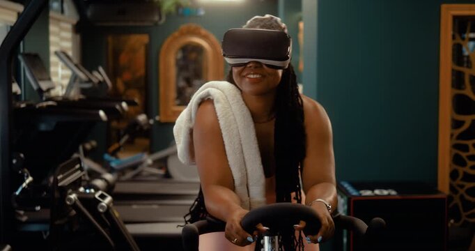 Excited Black woman enjoys a VR workout while riding a stationary bike in a modern gym, exploring a virtual world during cardio training.