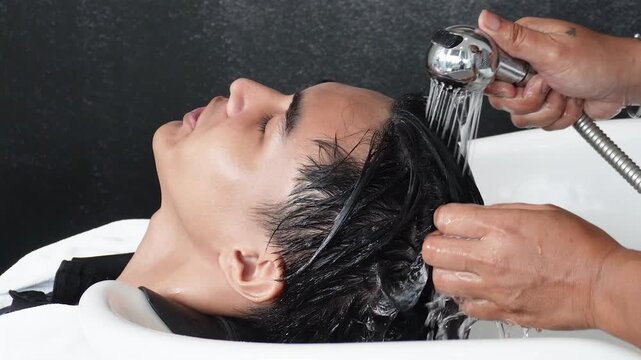 Hair stylist rinsing and washing young man's hair before professional haircut in salon for Southeast Asian man in his early 20s