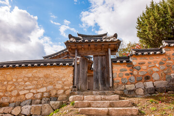 Fototapeta premium Exterior View of Traditional Korean Hanok Gate Featuring Stone Stairs, Stone Wall, Earthen Wall, and Wooden Door Under Blue Sky and Clouds