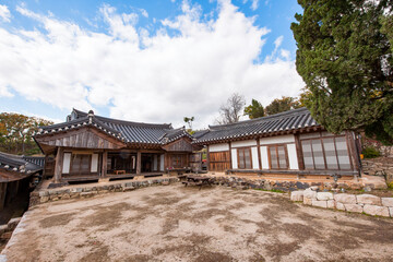 Obraz premium Exterior View of Traditional Korean Hanok Courtyard Featuring Wooden Buildings, Tiled Roofs, and Autumn Scenery Under a Cloudy Sky