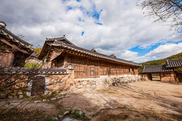 Obraz premium Exterior View of Traditional Korean Hanok House Featuring Stone Wall, Earthen Wall, Small Wooden Gate, and Tiled Roof Under a Cloudy Sky