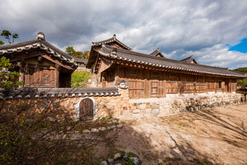 Fototapeta premium Exterior View of Traditional Korean Hanok House Featuring Stone Wall, Earthen Wall, Small Wooden Gate, and Tiled Roof Under a Cloudy Sky